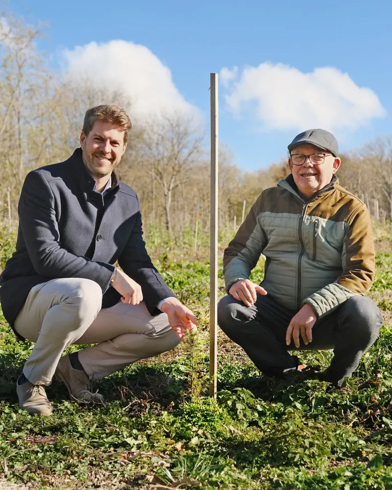 Two men are kneeling in a field with a wooden post between them. The man on the left is smiling and the man on the right is wearing a hat and glasses. They are surrounded by trees.