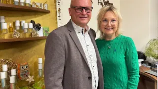 Ein Mann und eine Frau stehen lächelnd in einem Raum mit Regalen voller Flaschen und Dekorationsartikeln. Die Frau trägt einen grünen Pullover und der Mann hat eine Brille. Hinter ihnen steht ein Schild mit der Aufschrift 'Anne'.