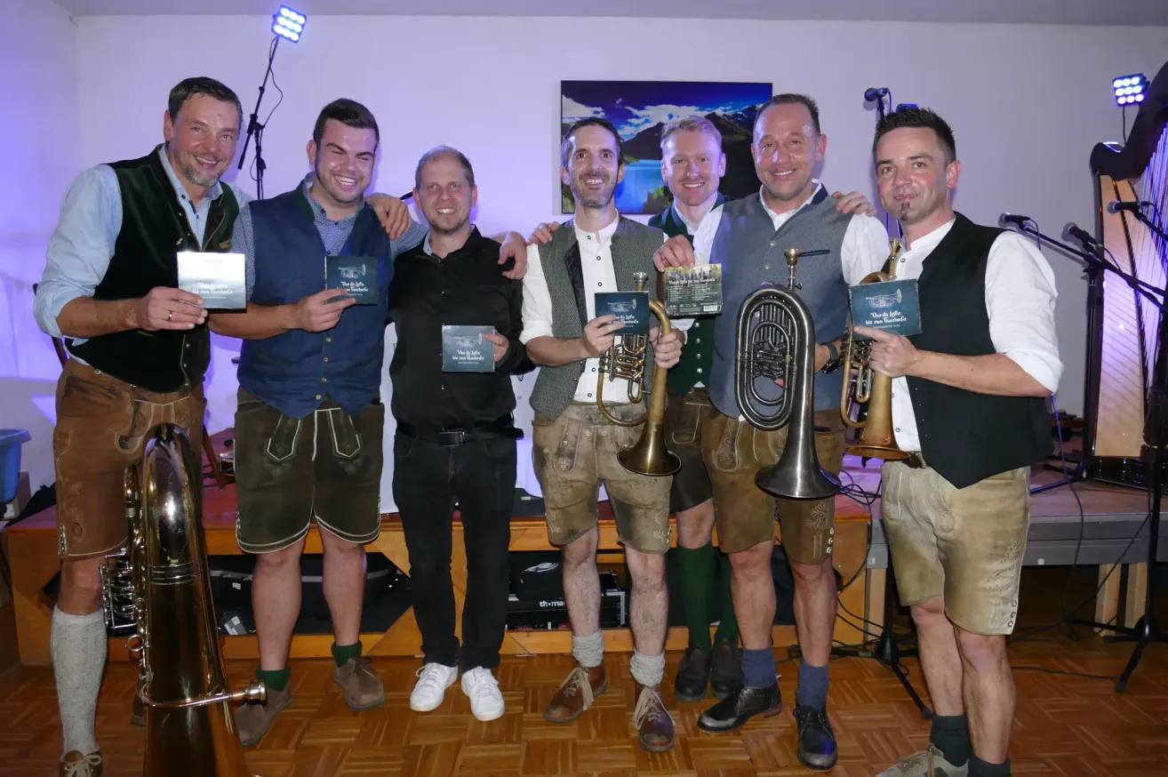 Sechs Männer in traditioneller Kleidung stehen lächelnd nebeneinander und halten Musikinstrumente und Auszeichnungen. Die Umgebung scheint eine Feier oder Preisverleihung zu sein, mit einem Hintergrundbild einer Landschaft.