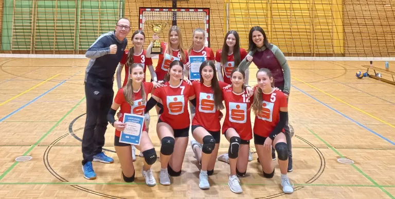A group of young women in red jerseys are posing for a photo on a court with a trophy, certificates, and a man standing behind them.