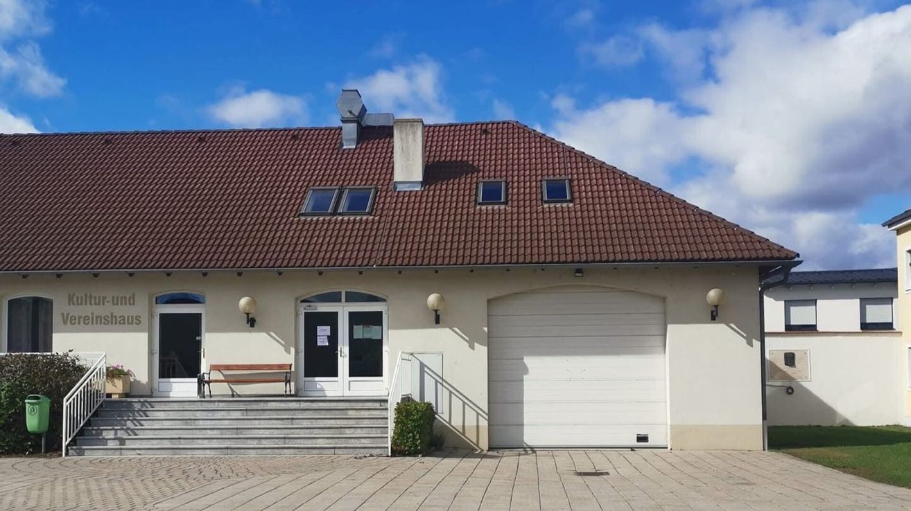 Ein Gebäude mit einem braunen Ziegeldach, mit einer einzelnen Bank, doppelten Glastüren, einer Garage und zwei Lampen auf jeder Seite.