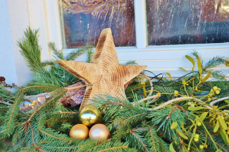 A large wooden star ornament is displayed on a bed of evergreen branches, with golden baubles and lights in the foreground, set against a rainy window backdrop.