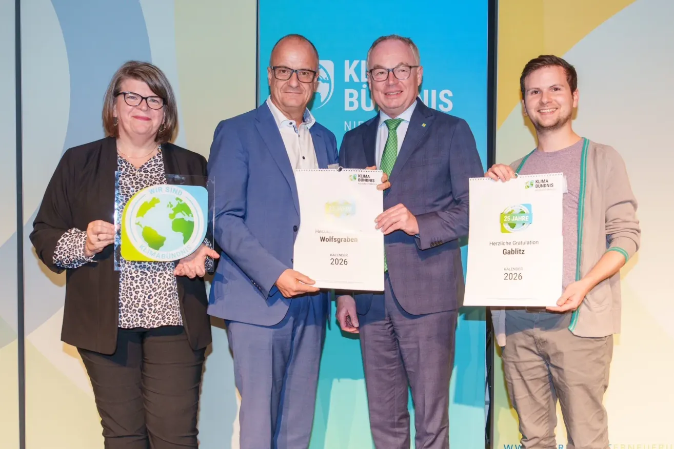 Four people stand together holding awards. Two men and a woman hold certificates. The men wear suits, the woman a dress. The background is a banner with 'Klima Bundnis' in large letters.