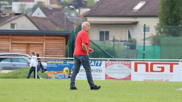 An older man walks on a grassy field with his hands in his pockets. Behind him, there are houses and a banner with text.