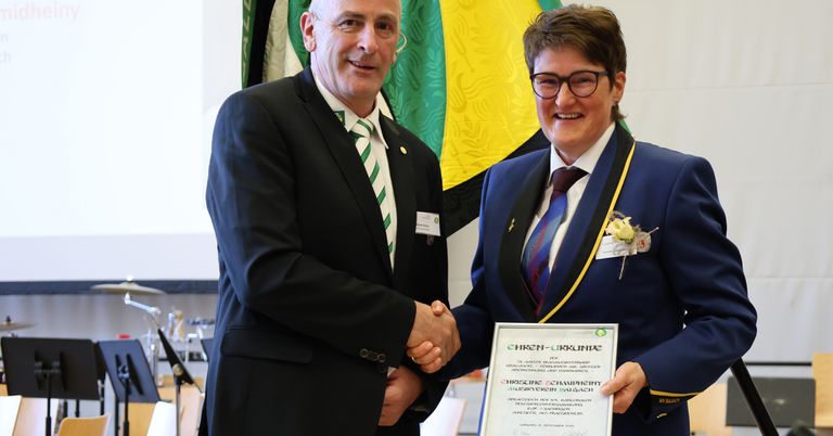 A man and a woman in formal attire are shaking hands in front of a flag, with the woman holding a certificate.