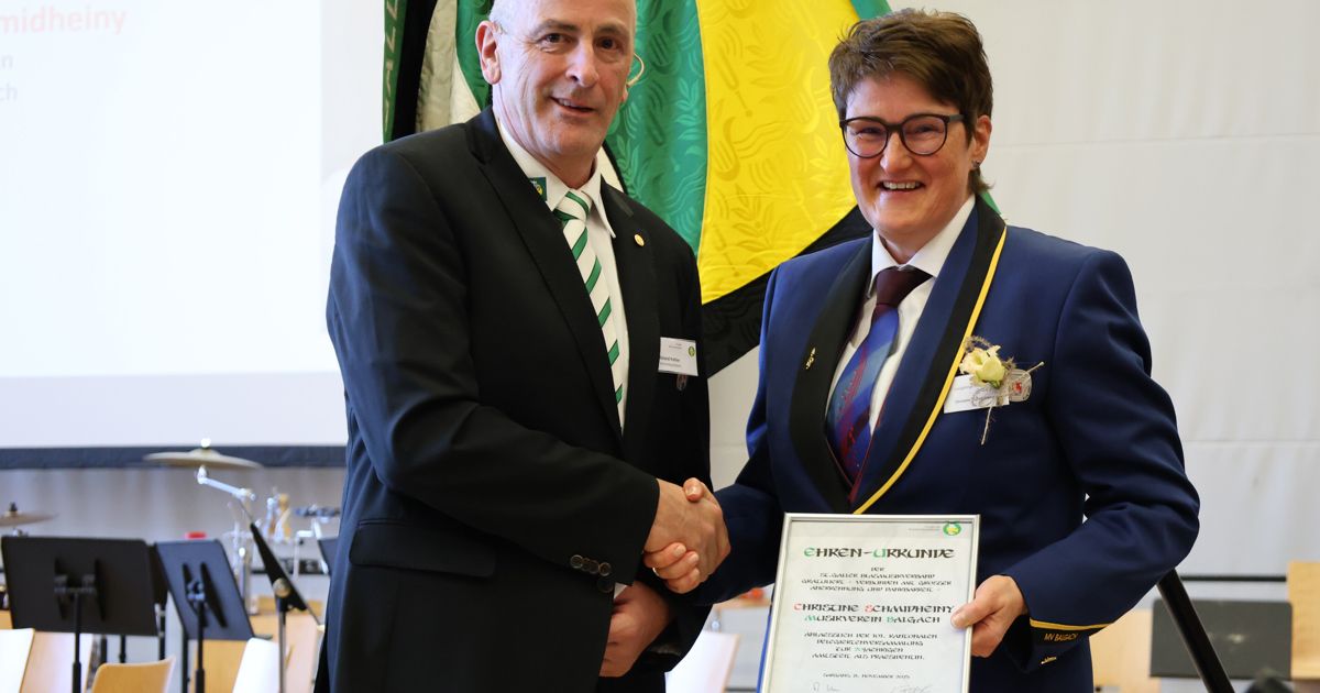 A man and a woman in formal attire are shaking hands in front of a flag, with the woman holding a certificate.
