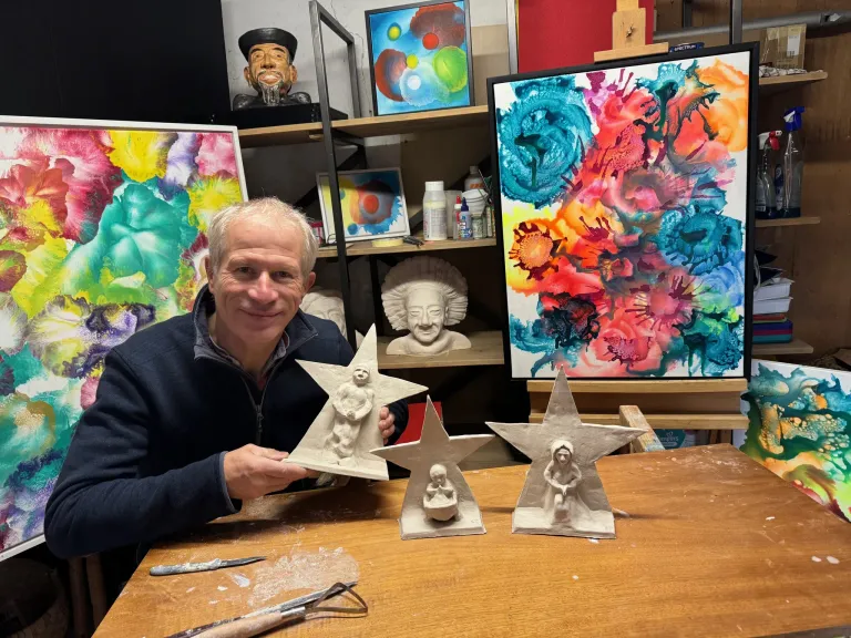 A man in a studio is smiling while holding two clay star sculptures. Behind him, various artworks and sculptures are displayed on shelves and the wall.