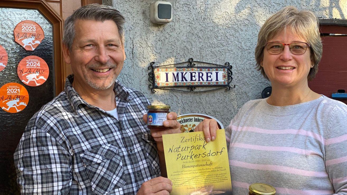 A couple standing in front of a wall with a sign reading 'IMKEREI'. The man holds a jar of honey and a certificate for 'Naturpark Purkersdorf'. They both smile.