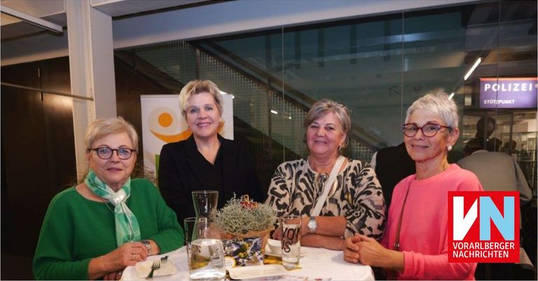 Four women are seated at a table, smiling. A flower arrangement and glasses are on the table. A staircase is visible in the background.