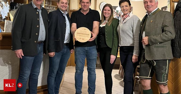 Eine Gruppe von Personen steht drinnen und posiert mit einer Plakette. Der Mann in der Mitte hält eine Holzplakette mit Logo und Text.