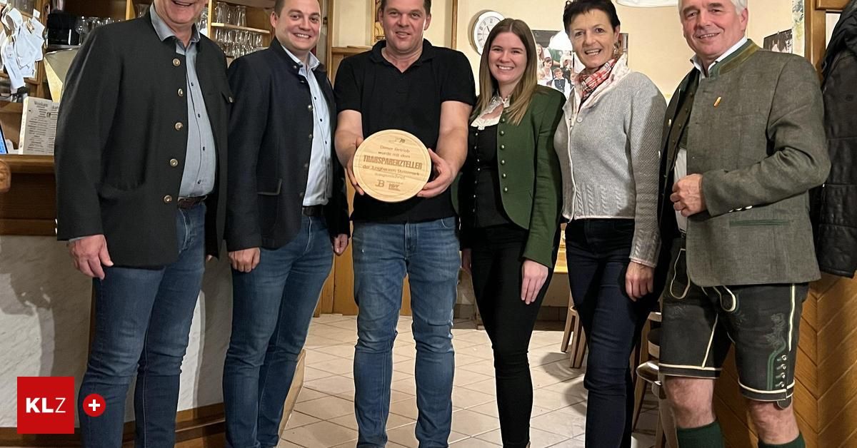 A group of individuals stand indoors posing with a plaque. The man in the center holds a wooden plaque with a logo and text.