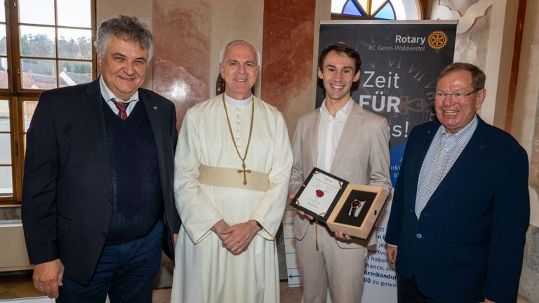 Vier Männer stehen lächelnd zusammen. Einer hält ein gerahmtes Dokument in einer Schachtel, und ein Priester steht neben ihm. Dahinter befindet sich ein Schild mit dem Text 'Zeit für'.