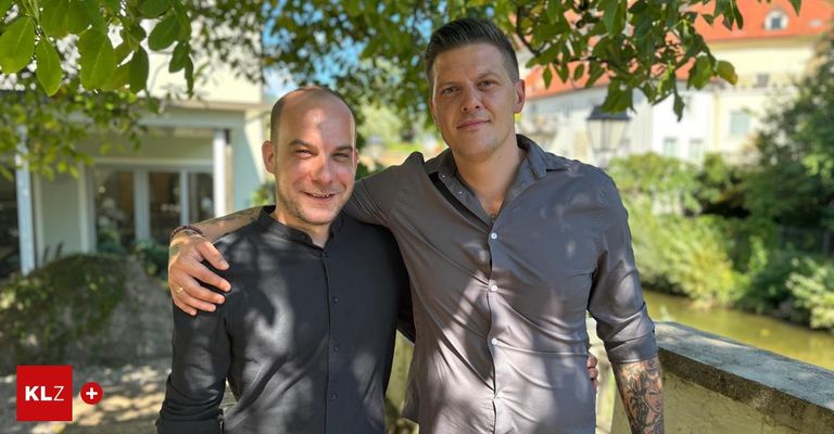 Two men standing close, one with a tattoo on his left arm, both smiling, under a tree. Behind them is a building and a bridge.