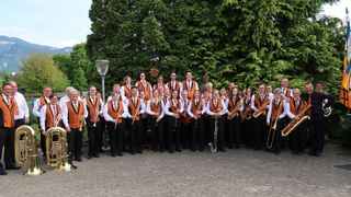 A large group of musicians in orange vests and black trousers are posing for a photo with their instruments in a park with trees and a street light.