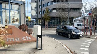 A black car drives past a street corner with a sign and a white box with the text 'a.ch' on a concrete base.