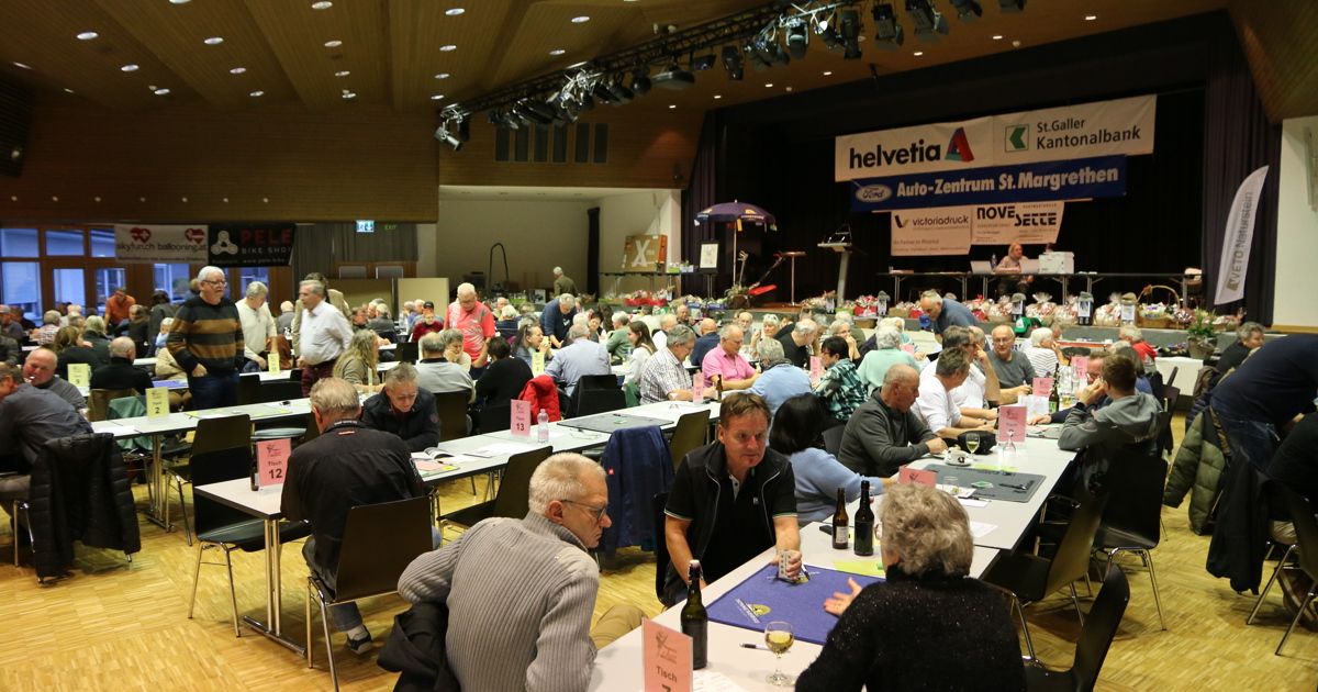 A large group of people are seated at tables in a room. Some have wine glasses, others have bottles. There are banners on the stage, and the room has a ceiling with lights.