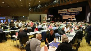 A large group of people are seated at tables in a room. Some have wine glasses, others have bottles. There are banners on the stage, and the room has a ceiling with lights.