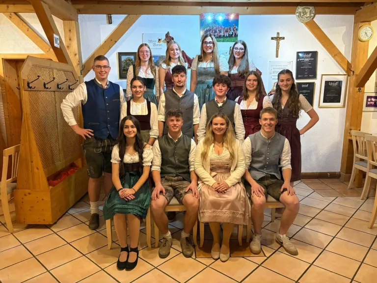A group of people dressed in traditional attire are posing for a photo in a room with wooden beams and tiled flooring. They are standing and sitting on chairs. There is a cross on the wall and some picture frames.
