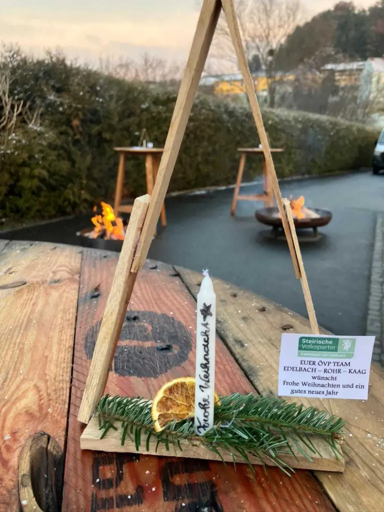 Ein dreieckiger Ständer mit einer Kerze, Tannennadeln und einer Orangenscheibe. Dahinter zwei Holztische mit Feuerstellen und ein Schild, das 'Edelbach - Rohr - Kaag Frohe Weihnachten und ein gutes neues Jahr' sagt.