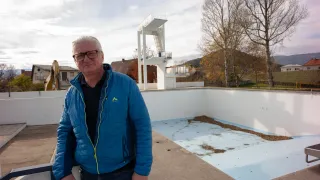 A man wearing glasses and a blue jacket stands next to an empty pool with a diving tower.