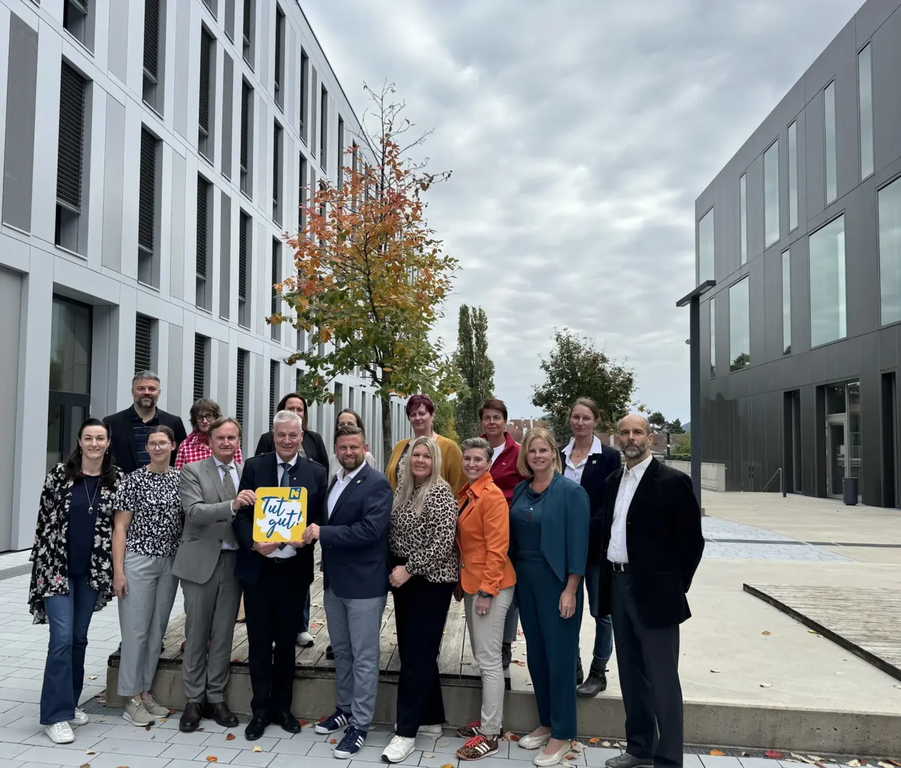 A group of individuals stands in front of a modern building. They are smiling and posing for a photo. A man holds a yellow sign with the words Tut gut!