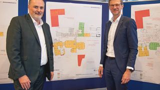 Two men are standing next to a large poster in an office. They are wearing suits and eyeglasses. The poster has a floor plan on it.