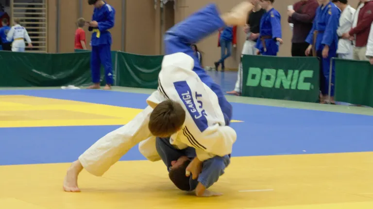 Two individuals are participating in a martial arts competition. One is executing a move while the other is on the floor. Spectators and participants are wearing blue uniforms.