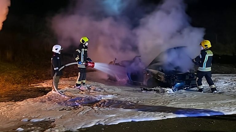 Two firefighters in protective gear are spraying water on a burning car emitting smoke. The scene is at night.