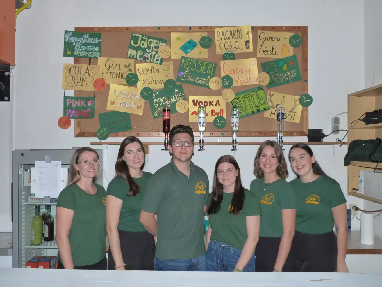 Eine Gruppe von Menschen in grünen Shirts posiert für ein Foto vor einem Korkbrett mit verschiedenen Getränken und einer Tafel mit aufgelisteten Getränken.