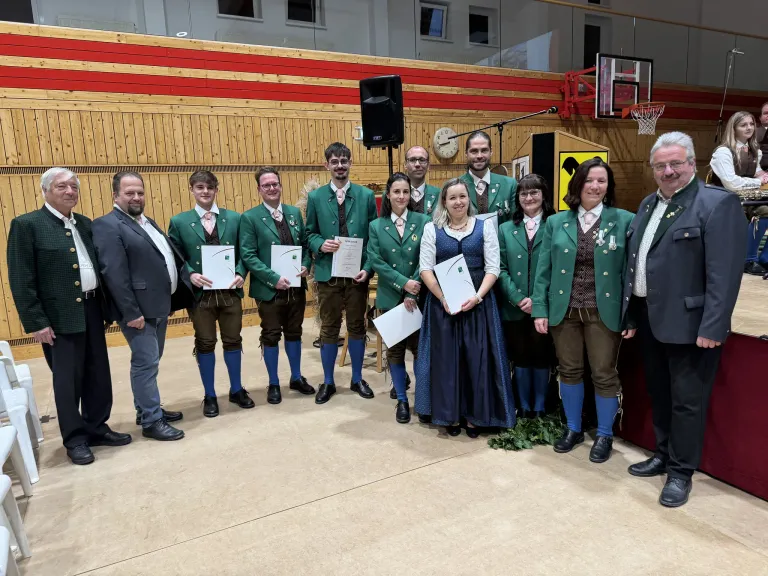 Eine Gruppe von Menschen in grünen Jacken und traditioneller Kleidung posiert für ein Foto in einer Turnhalle, mit einem Basketballkorb im Hintergrund.