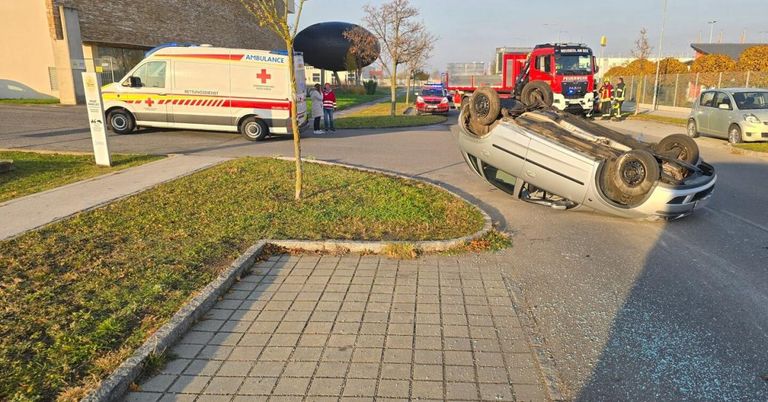 Ein Krankenwagen ist am Straßenrand geparkt. Ein Auto ist in der Nähe umgekippt. Zwei Personen stehen in der Nähe des Krankenwagens. Rettungsfahrzeuge sind am Unfallort.