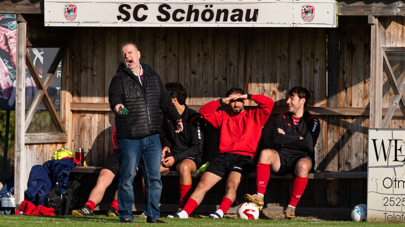 Ein Fußballtrainer schreit Spieler auf der Bank an, einer bedeckt seine Augen, ein anderer lächelt und ein Fußball liegt in der Nähe.