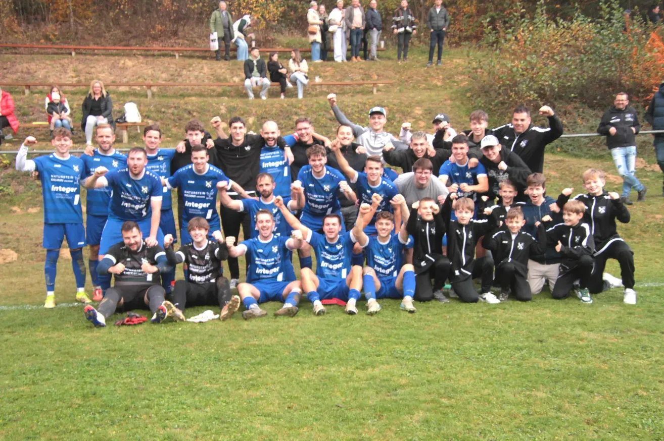 Eine Gruppe von Fußballspielern in blauen Trikots posiert für ein Mannschaftsfoto auf einem Rasenfeld, mit mehreren Personen im Hintergrund, die stehen und sitzen.