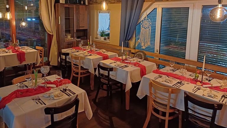 Ein Restaurant mit Holzstühlen und für eine Mahlzeit gedeckten Tischen. Die Tische sind mit weißen Tischdecken, roten Servietten und Besteck geschmückt. Der Raum hat große Fenster, einen Holzschrank und einen hängenden Traumfänger.