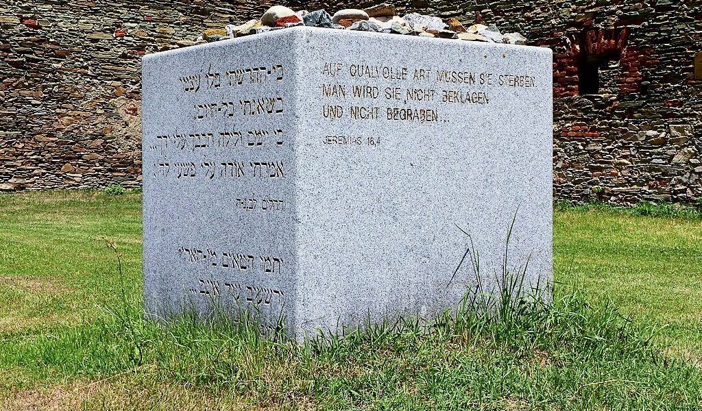 Ein Grabstein auf einem Friedhof mit hebräischen und deutschen Inschriften. Er steht auf Gras mit Steinen darüber. Dahinter ist eine Ziegelmauer.