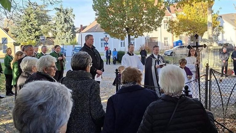 Mehrere Menschen versammeln sich im Freien in der Nähe eines Baumes, wahrscheinlich für eine Veranstaltung. Ein Priester liest aus einem Buch, und andere hören zu. Ein Gebäude und Autos sind im Hintergrund.