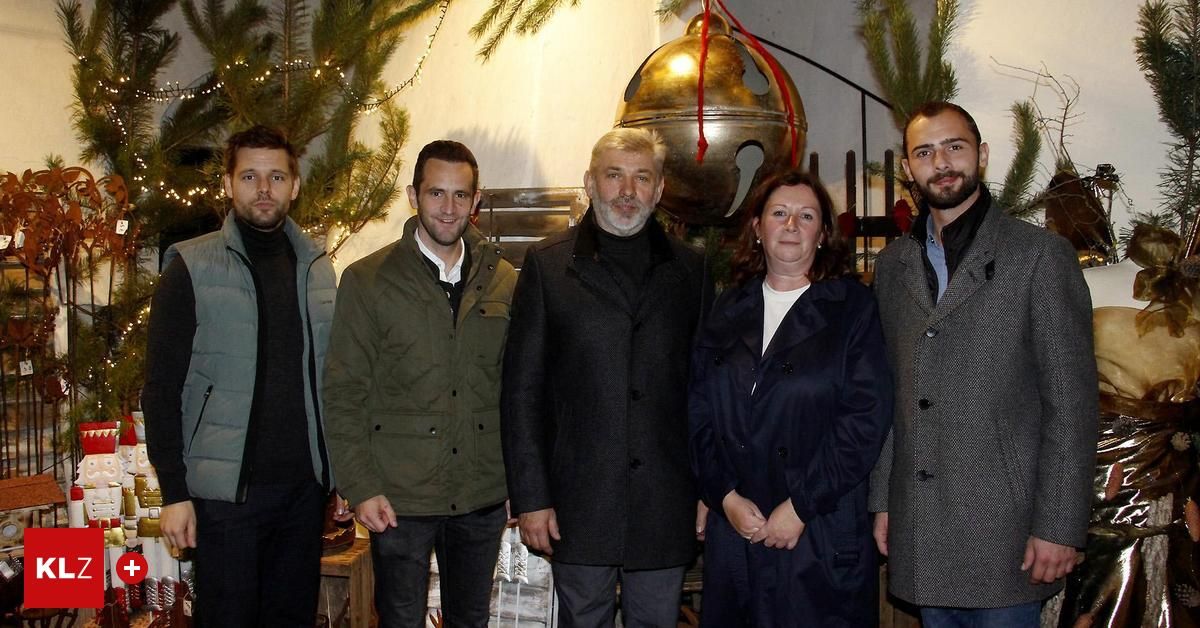 Fünf Personen, darunter zwei Frauen und drei Männer, stehen eng beieinander in einer festlichen Umgebung und lächeln für ein Foto. Eine große Glocke hängt hinter ihnen.