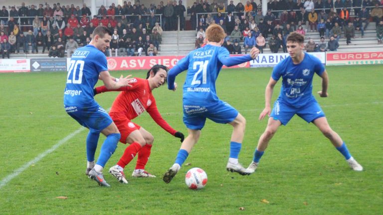 Ein Fußballspiel mit Spielern in blauen und roten Trikots. Der Spieler in Blau, Nummer 21, tritt den Ball. Ein Spieler in Rot versucht, ihn zu blockieren.