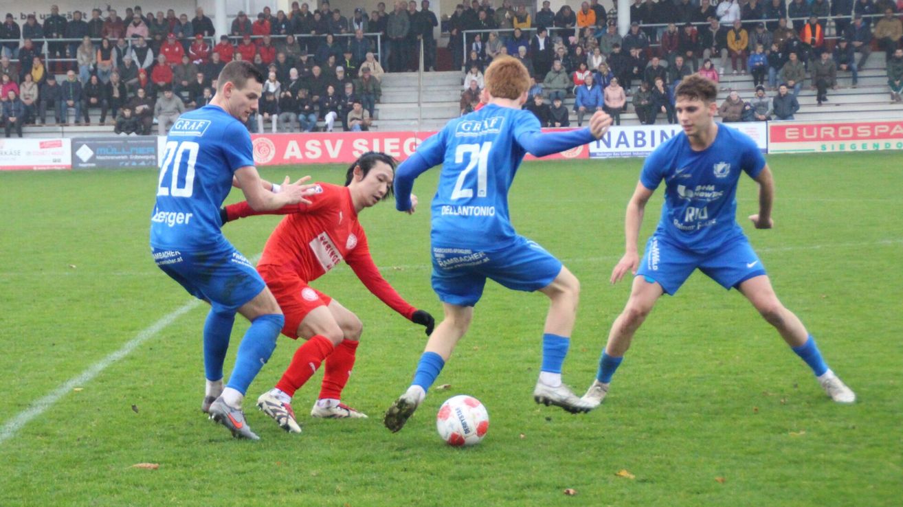 Ein Fußballspiel mit Spielern in blauen und roten Trikots. Der Spieler in Blau, Nummer 21, tritt den Ball. Ein Spieler in Rot versucht, ihn zu blockieren.