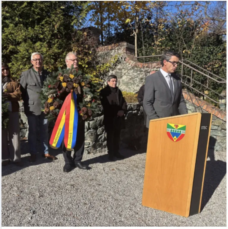A man holding a wreath stands at a podium with a coat of arms, while others stand behind him in a park.