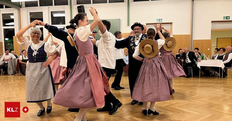 Eine Gruppe von Menschen tanzt in einem Saal. Sie tragen traditionelle Kleidung. Ein Mann in einem Hut führt den Tanz an. Es gibt Menschen, die von den Seiten zuschauen.