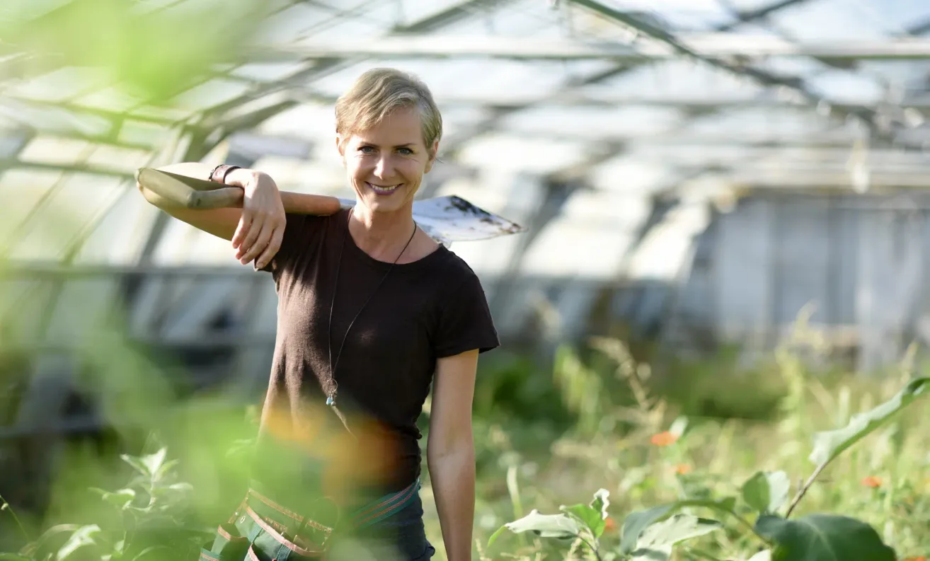 Eine Frau im Gewächshaus, die eine Schaufel hält, ein braunes T-Shirt und eine Halskette trägt, mit einem unscharfen Hintergrund von Pflanzen und einer Gewächshausstruktur.