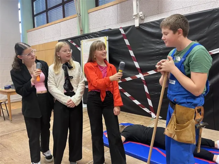 Vier Personen stehen in einem Raum, zwei mit Mikrofonen, eine weitere hält einen Stock und eine Person liegt auf einer blauen Matte.