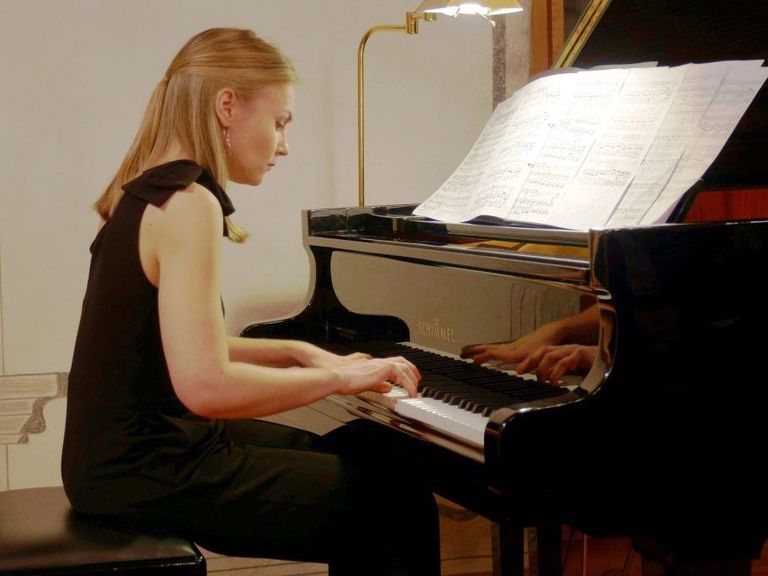 Eine Frau sitzt an einem Klavier, konzentriert auf das Notenblatt vor ihr, beleuchtet von einer Lampe. Sie trägt ein schwarzes ärmelloses Oberteil und Hose.