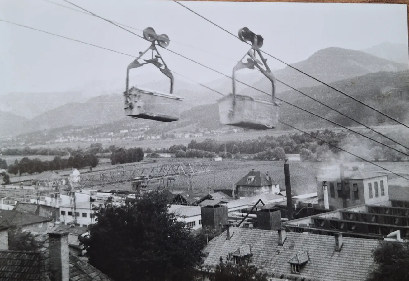 Black and white photo of two cable cars in the air with a town below, mountains in the distance, and a factory emitting smoke.