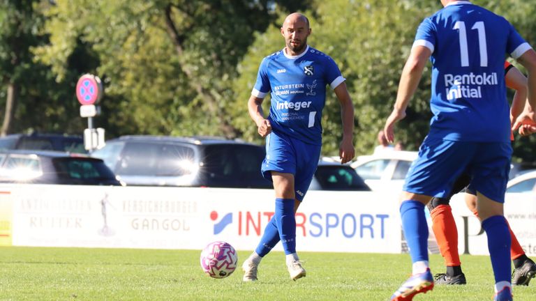 Ein Fußballspieler in einem blauen Trikot mit 'Integrer' auf der Vorderseite läuft auf dem Feld. Hinter ihm ist ein weißer Banner mit 'Gangol' und 'INTRSPORT' zu sehen. Im Hintergrund sind Autos geparkt.