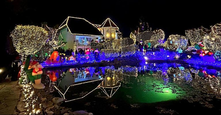 Ein Haus, das mit bunten Lichtern beleuchtet ist, für die Feiertage geschmückt, mit einem Teich, der die Lichter reflektiert, und Menschen, die die festliche Dekoration genießen.