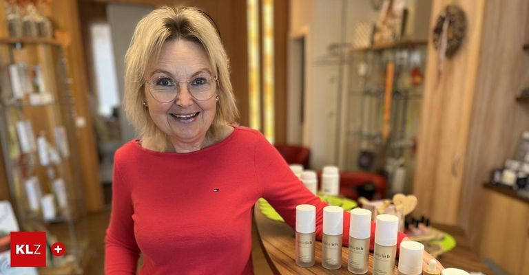 Eine Frau mit blonden Haaren, die eine Brille und ein rotes Langarmshirt trägt, steht hinter einem Tresen mit mehreren weißen Flaschen von Hautpflegeprodukten.