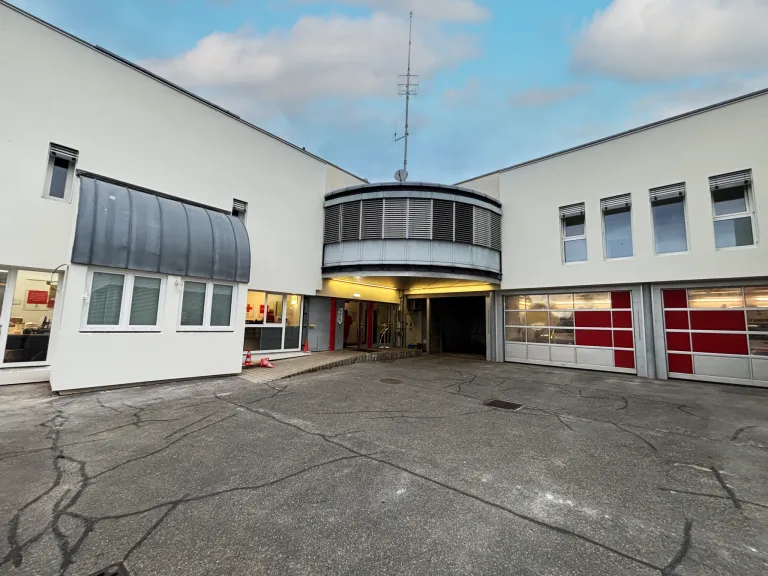 Ein modernes Gebäude mit einem geschwungenen Eingang, Fenstern und einem grauen Vordach. Ein klarer blauer Himmel mit Wolken ist darüber. Das Gebäude hat eine Garage mit roten und weißen Türen.