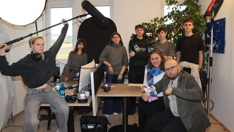 Eine Gruppe von Menschen befindet sich in einem Studioraum. Ein Mann hält ein Mikrofon. Eine Frau hält ein Bild. Eine Frau sitzt an einem Tisch mit einer Flasche, einer Schachtel und einer Kamera.